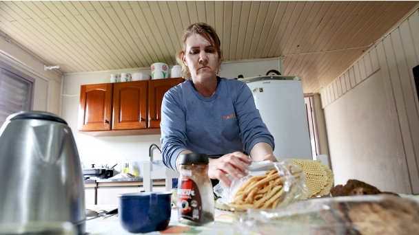 A foto mostra uma mulher em uma cozinha. Ao fundo, há armários marrons e uma geladeira branca. Sobre a mesa, estão dispostos utensílios e alimentos.