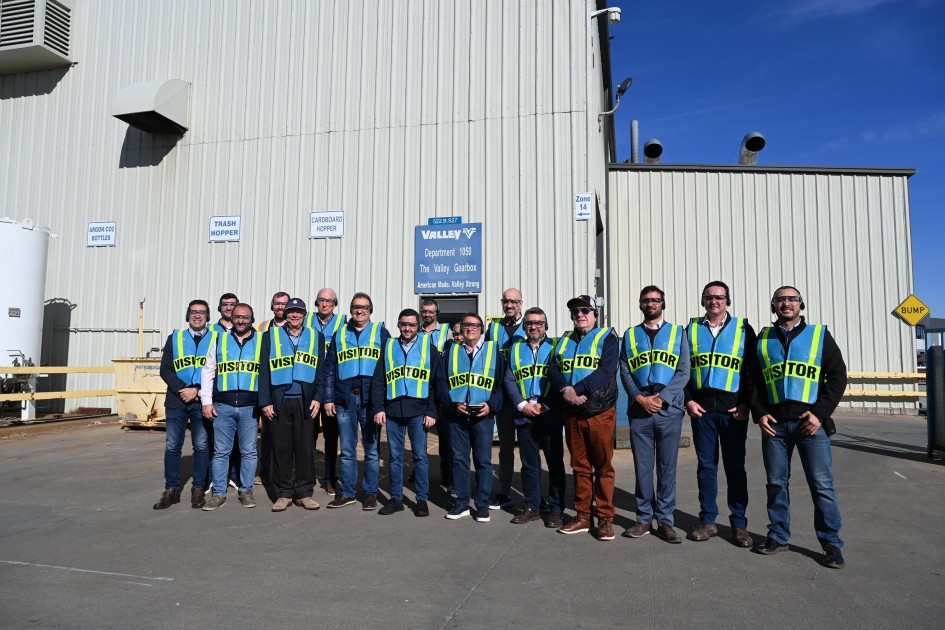 A imagem apresenta um grupo de visitantes posicionado em frente a um grande galpão industrial de paredes metálicas claras. Todos vestem coletes de segurança fluorescentes com a inscrição “VISITOR”.