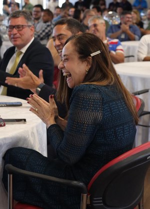 Audiodescrição: Imagem de um grupo de pessoas sentadas. Em destaque, uma mulher que está sorrindo 