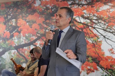 Audiodescrição: Imagem de homem falando ao microfone. Ele usa termo na cor cinza