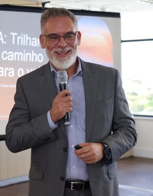 Audiodescrição: Imagem de um homem falando ao microfone. Ele usa óculos, tem barba e cabelo grisalhos 