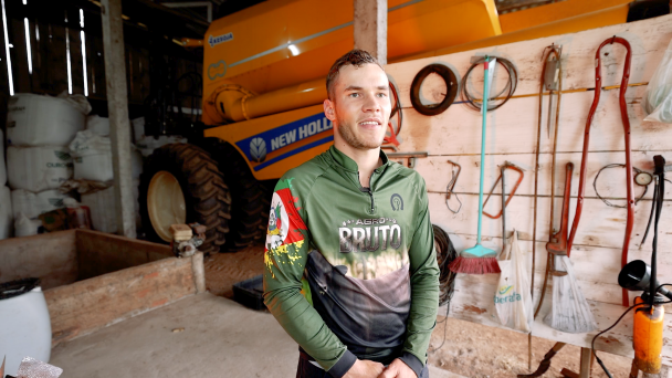 A imagem apresenta um homem em pé dentro de um galpão agrícola. Ao fundo, há uma grande máquina agrícola. Na parede lateral, estão pendurados diversos utensílios e ferramentas.