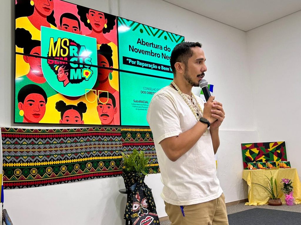 Um homem de camisa branca e colares fala ao microfone, em frente a um telão colorido com o tema "Abertura do Novembro Negro" e a arte da campanha "MS Sem Racismo".
