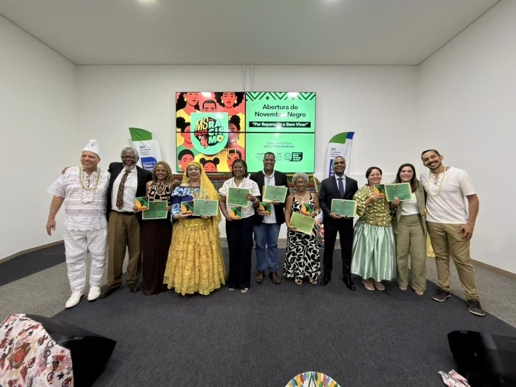 Grupo de homenageados e autoridades posa no palco com certificados. O grupo inclui pessoas em trajes sociais e tradicionais afro-brasileiros.
