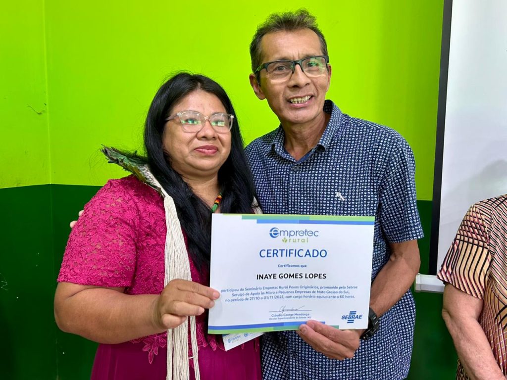 Uma mulher indígena, Inaye Gomes Lopes, usando um vestido rosa e um adorno com penas, sorri enquanto segura seu certificado do "Empretec Rural Povos Originários". Ao seu lado, um homem sorri. O certificado é do SEBRAE e indica a participação de 27/10 a 01/11/2023.