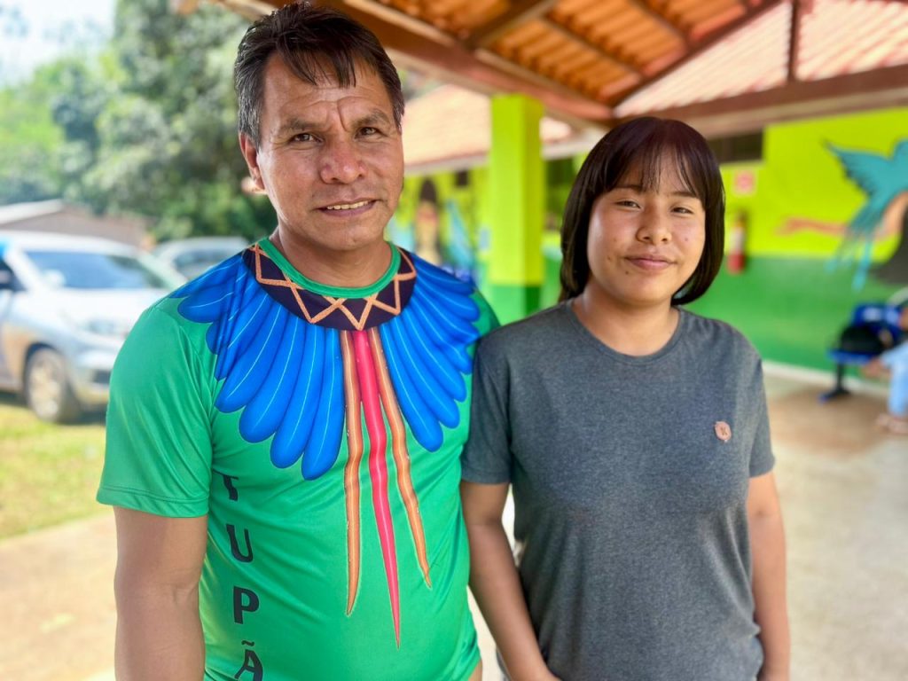 Um homem e uma jovem posam juntos, sorrindo. O homem veste uma camiseta verde com grafismos indígenas em vermelho e azul, e a palavra "TUPÃ" na frente. A jovem está ao seu lado, vestindo uma camiseta cinza simples.