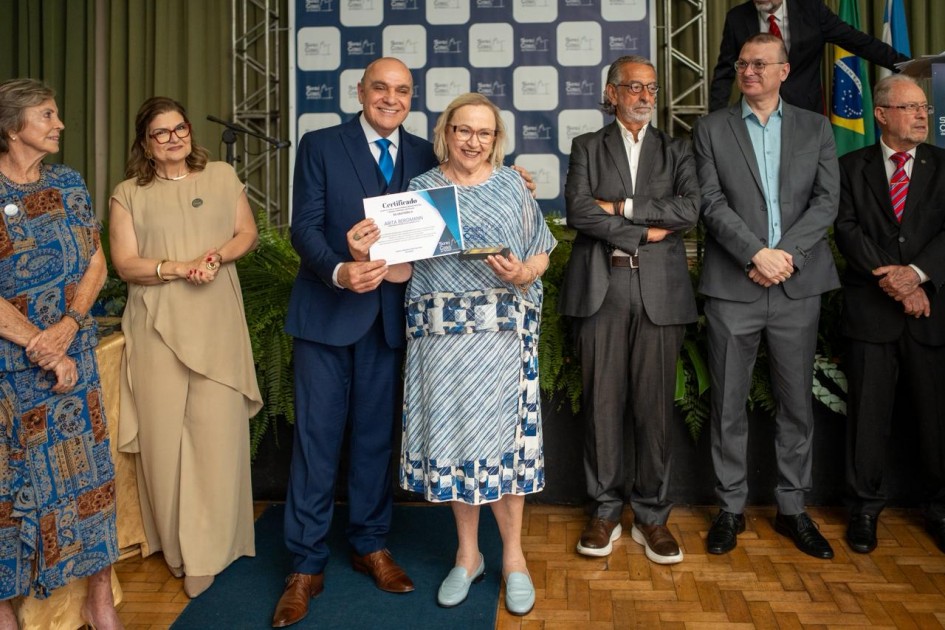 A imagem registra uma cerimônia oficial de entrega de certificados na Santa Casa de Pelotas. Ao centro da foto, uma mulher de cabelos loiros e óculos — Arita Bergmann, secretária da Saúde do Rio Grande do Sul — aparece sorrindo enquanto recebe um certificado e segura também uma pequena estatueta.
Ao lado dela está um homem de terno azul escuro, que entrega o certificado. À direita e à esquerda, demais autoridades acompanham a homenagem posicionadas em fila. O fundo exibe um painel institucional azul e branco do evento.