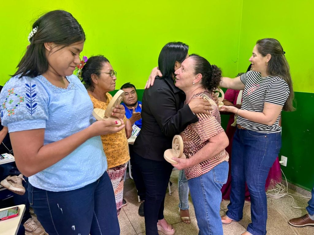 Momento de abraço e celebração. Uma mulher de preto abraça outra mulher com uma blusa listrada bege e preta. Mulheres indígenas ao redor seguram e admiram pequenos objetos de madeira entalhados, que são troféus do evento.