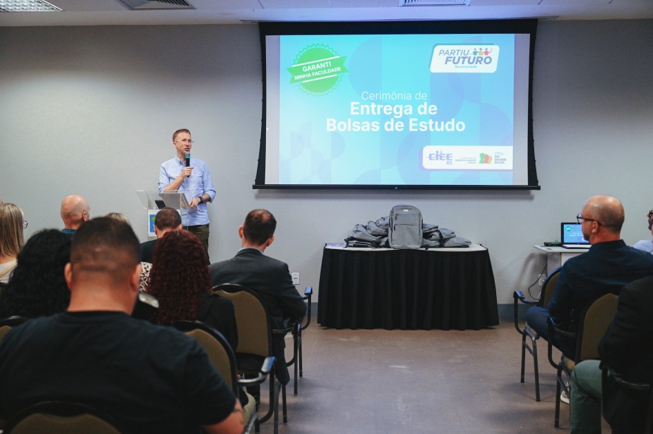 Foto do fundo de um auditório, enquadrando Fantinel falando ao microfone no púlpito. Há uma tela no fundo com a exibição de um slide. Várias pessoas aparecem assistindo à palestra, sentadas.