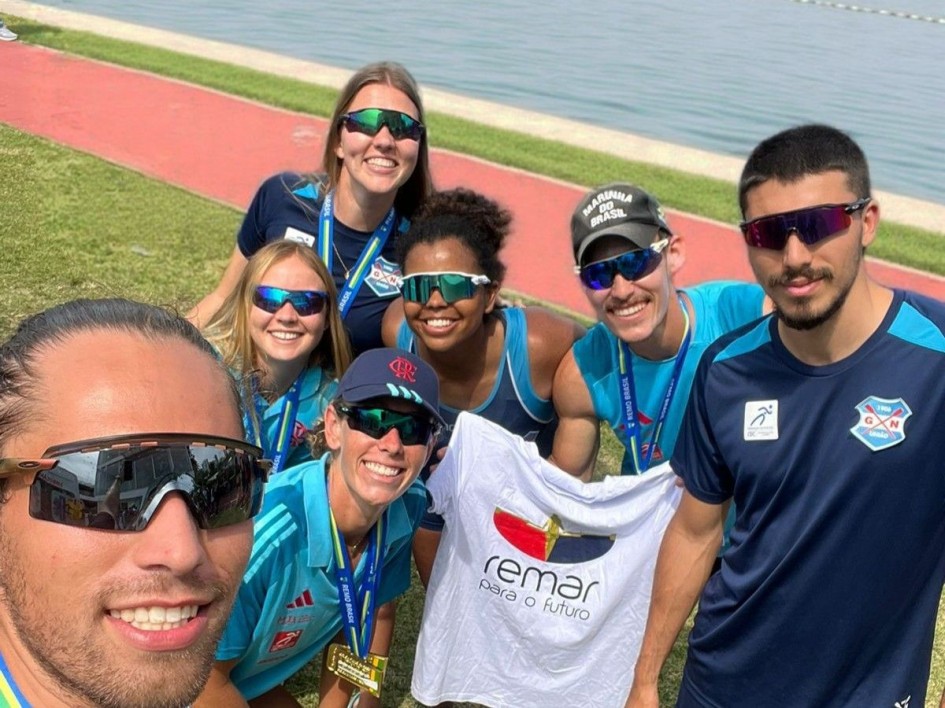 Um grupo de sete pessoas posa para uma selfie ao ar livre, próximo a um lago. Todos usam óculos esportivos escuros e roupas de treino. Alguns usam medalhas no pescoço. Uma das pessoas segura uma camiseta branca com o logotipo “Remar para o Futuro”. O fundo mostra área gramada e pista de concreto ao lado da água.