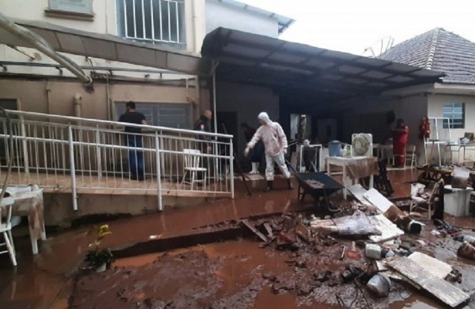 A imagem mostra a área externa de um hospital após uma enxurrada. O chão está coberto de lama espessa e detritos espalhados, como móveis, utensílios e pedaços de estruturas danificadas. Algumas pessoas trabalham na limpeza, usando pás e equipamentos improvisados. Parte da estrutura do prédio apresenta danos e acúmulo de sujeira nas áreas inferiores.