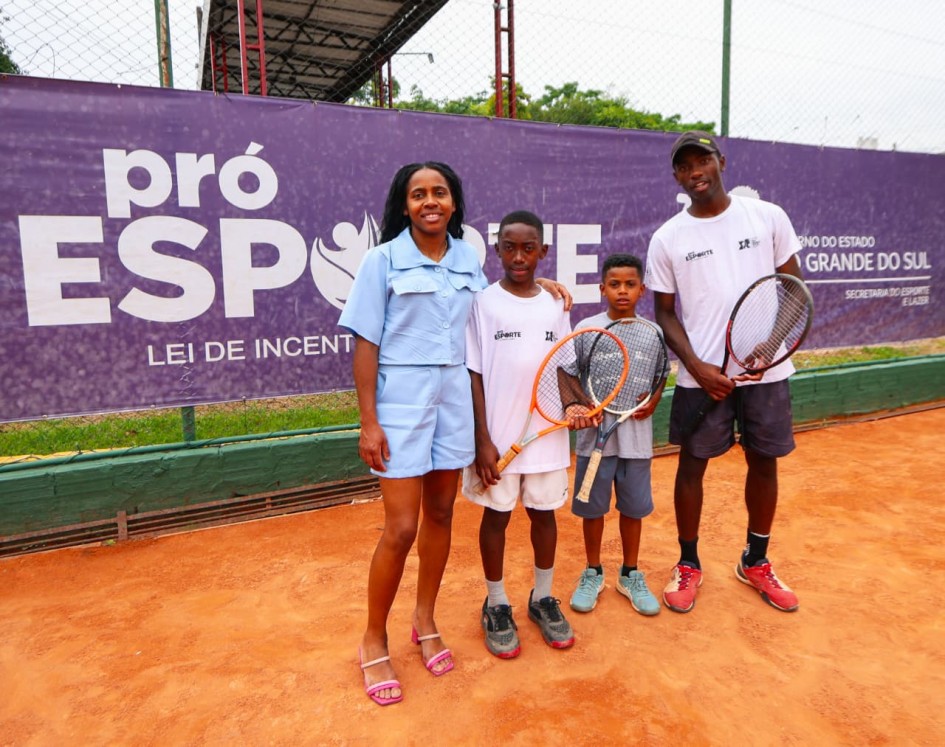 Quatro pessoas estão em uma quadra de saibro de tênis, diante de um grande banner roxo com o texto “Pró-Esporte – Lei de Incentivo ao Esporte”. À esquerda, uma mulher veste conjunto azul claro. Ao lado dela, duas crianças seguram raquetes de tênis. O adulto à direita veste camiseta esportiva branca e segura uma raquete. Todos estão próximos à rede de tênis e o ambiente ao redor mostra vegetação e arquibancadas.